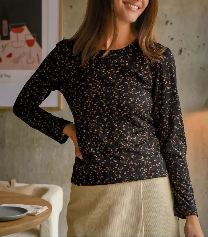 A woman standing in a room with a black floral top and beige skirt, smiling at the camera.
