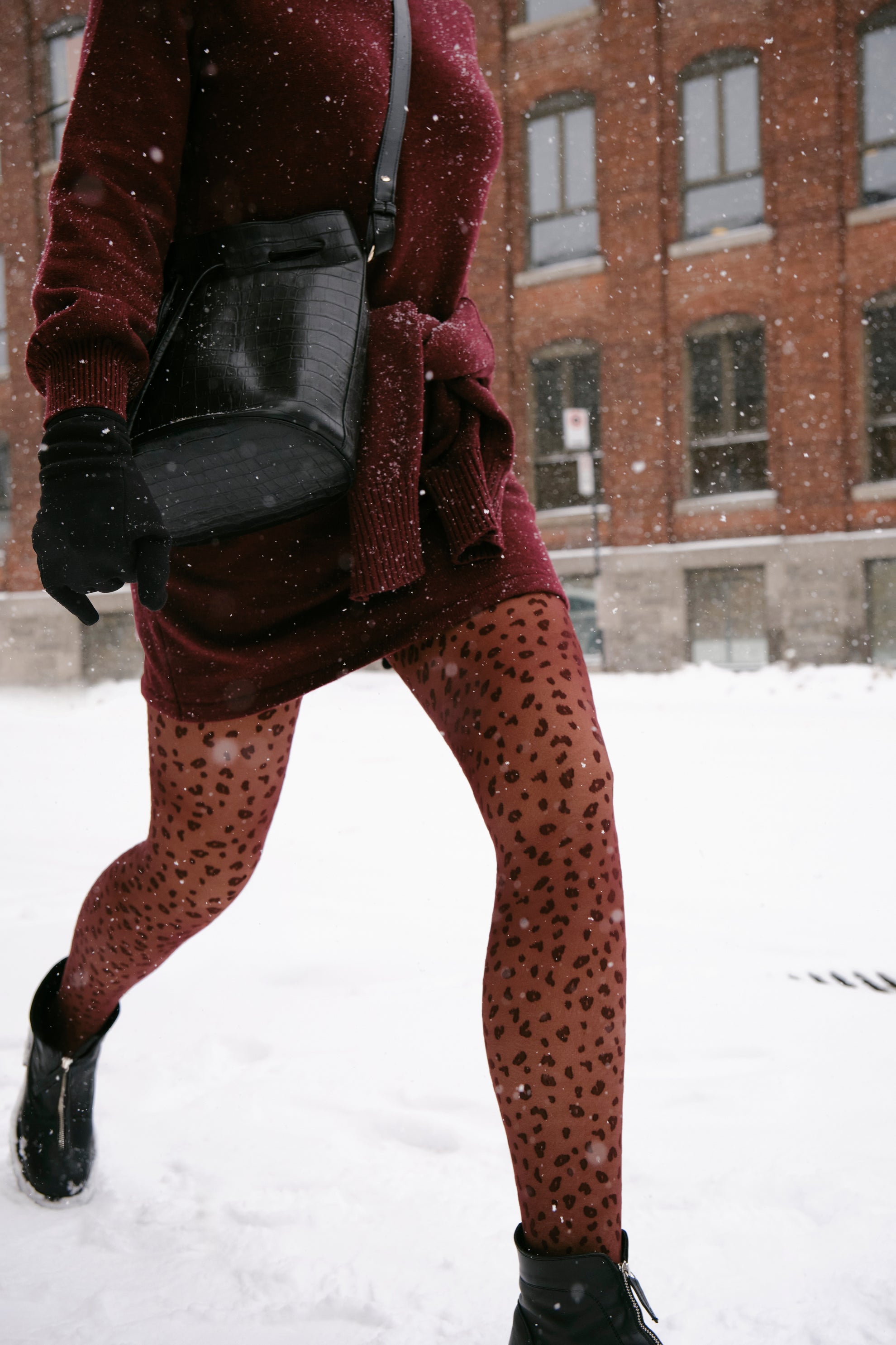 Person wearing a burgundy dress, black gloves, and leopard patterned tights walking in the snow.
